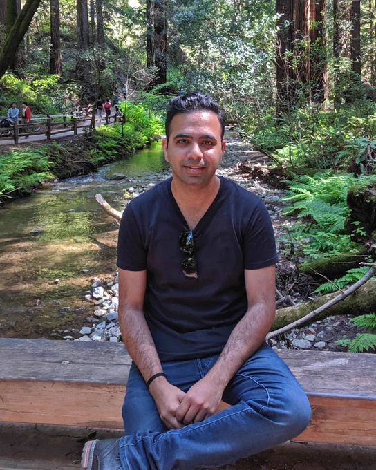 Sameer smiling sitting on a rock in front of water and wearing blue jeans and a black long sleeve shirt.