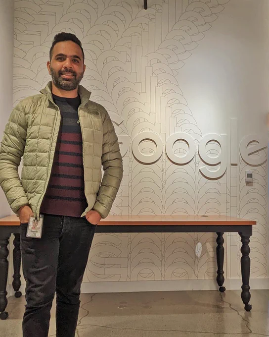 Sameer smiling standing in front of white Google sign wearing a tan jacket, red striped shirt, and black jeans. 