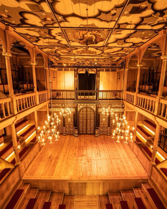 A view from the Upper Gallery of the Sam Wanamaker Playhouse looking down onto an empty theatre and stage. Wooden benches with red cushions are arranged in a horseshoe in front of a wooden thrust stage. Six lit candelabra hang from the ceiling, with tapered candles shining golden light onto the space.
