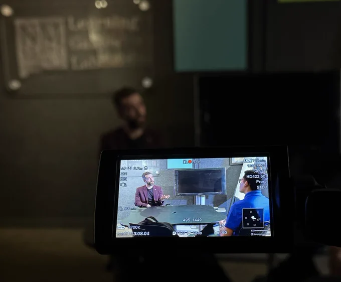 The foreground shows a bright display from a video camera of two men talking at a table. In the background the same two people are shown deeply shadowed.