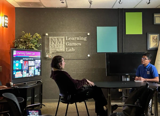 A video display and a sign each read "NM State University Learning Games Lab." Next to these signs, two men are sitting and talking at a small table. 