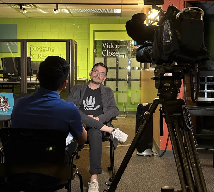 Two men sit and talk, with a video camera shown in the foreground taking footage of their conversation. In the background, glass doors and partitions read "blogging and research" and "video closet."