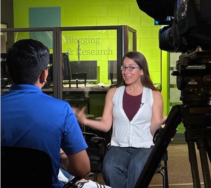 A woman speaks in front of the camera. Behind her a glass partition reads "Blogging and Research." The man interviewing her can also be seen from behind.