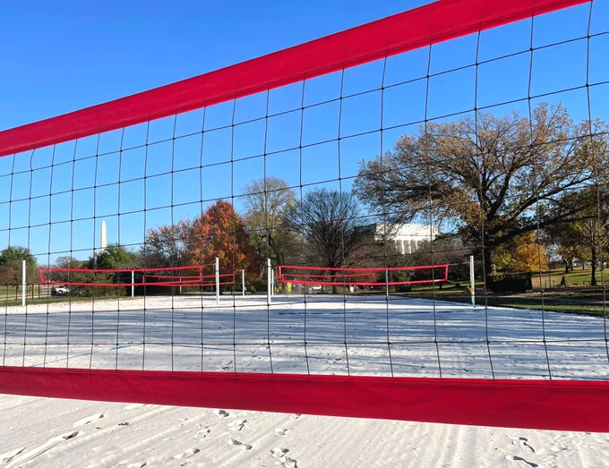 Looking through a volleyball net at several other sand volleyball courts in a park surrounded by trees with the Lincoln Memorial and Washington Monument standing in the distance.