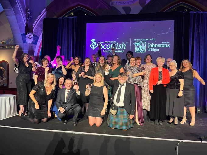Group of people standing on a stage holding Scottish Thistle Awards. Screen in background with text Scottish Thistle Awards with Johnstons of Elgin logo.