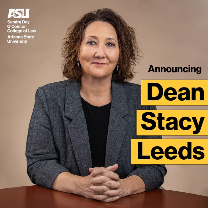 Photo of a smiling woman seated at a table in front of a brown backdrop. She has shoulder-length wavy brown hair, light brown complexion, wearing a black shirt with grey textured blazer. Text: “Announcing” at top. Beneath that, “Dean Stacy Leeds”
