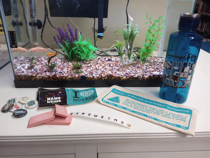 Fishtank on a counter with NaNoWriMo themed buttons, stickers, and waterbottle.