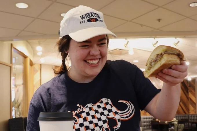 a smiling Bon Appetit student employee holding up a sandwich wearing a Wheaton College hat and screenprinted gray shirt