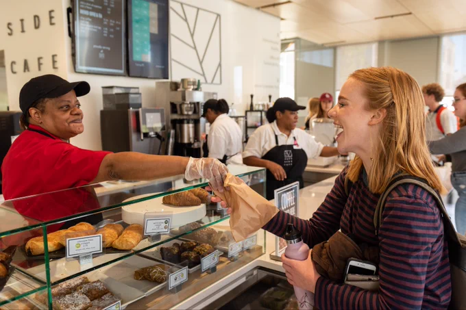 a smiling Bon Appetit employee handing a smiling guest on their way to class a pastry in a paper bag