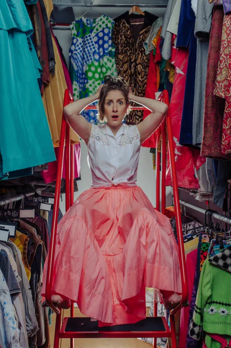 Photograph of a woman in a large closet, sat on a red set of steps. She is surrounded by brightly coloured clothing and is wearing a large, pink skirt and white blouse. She is holding her hands to her head and looking at the camera with her mouth wide open.