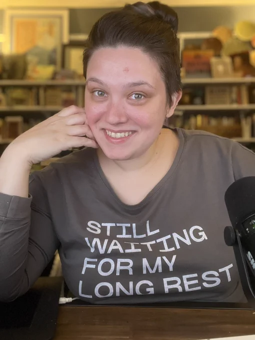 Emily Friedman smiles at camera. She is wearing a gray sweatshirt that reads “still waiting for my long rest” in white letters.