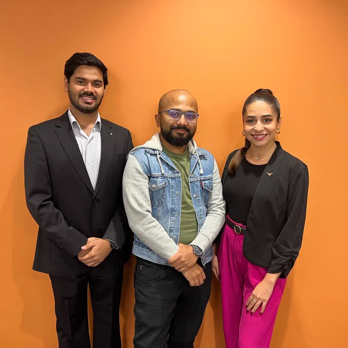 Two men and a woman smiling and standing together in front of an orange wall