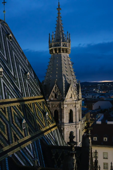 A photo of a small tower on the Votivkirche. the roof of the main building is on the right and has a pattern of black, white, green, and yellow tiles forming chevrons and diamonds. The tower is in a neo-gothic style with many small protrusions from its roof as it slopes upwards. Clouds can be seen in the background, and the city sprawls out below.