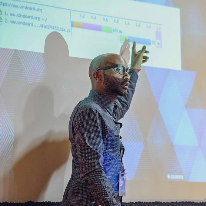 Person in glasses and a navy blue button up polkadot shirt, presenting at a conference, pointing at the screen.