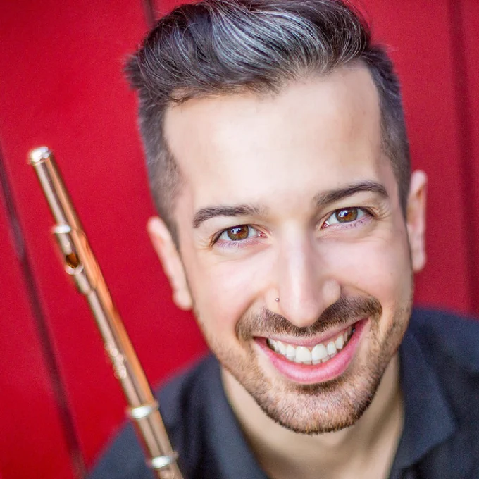 A close up head shot of flautist Christian Paquette, smiling at the camera and holding his flute