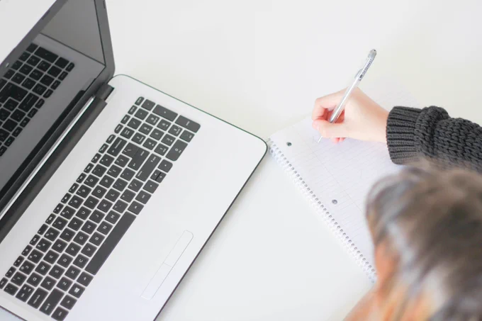 A child sitting in front of a laptop, using a notebook and a pen to write notes