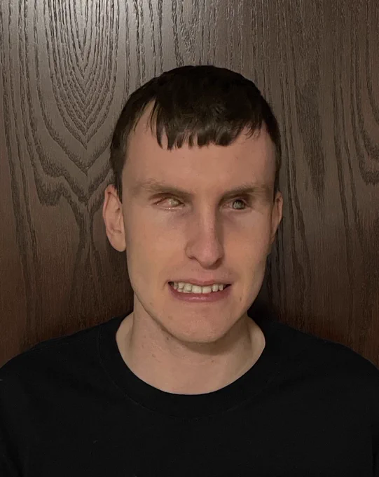 This photo shows Mitchell standing in a black shirt against a wooden background. He is smiling and looking at the camera. He has brown hair.