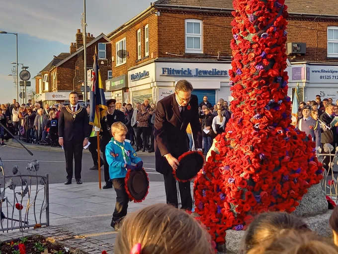 A Beaver Scout and an adult each lay a wreath in front of a war memorial covered in knitted poppies