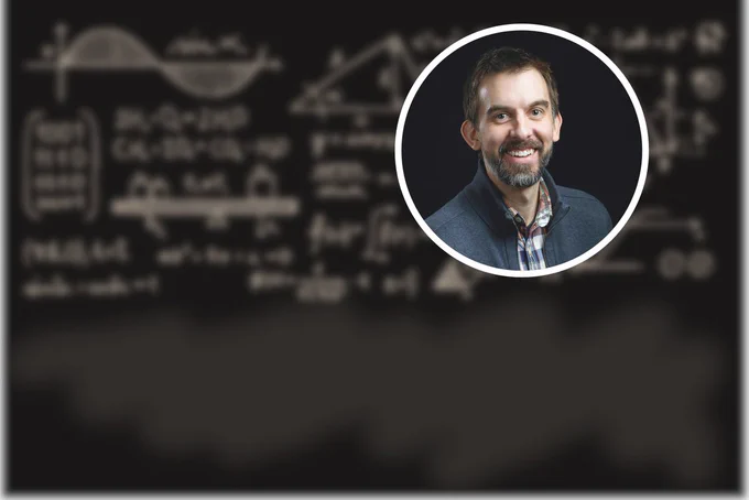 Headshot photo of James McKinlay (a white man in a suit jacket) in front of a blackboard background with blurred equations