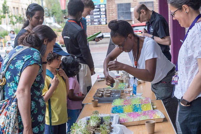 An adult supervises and smiles as children engage in arts and crafts outside a library