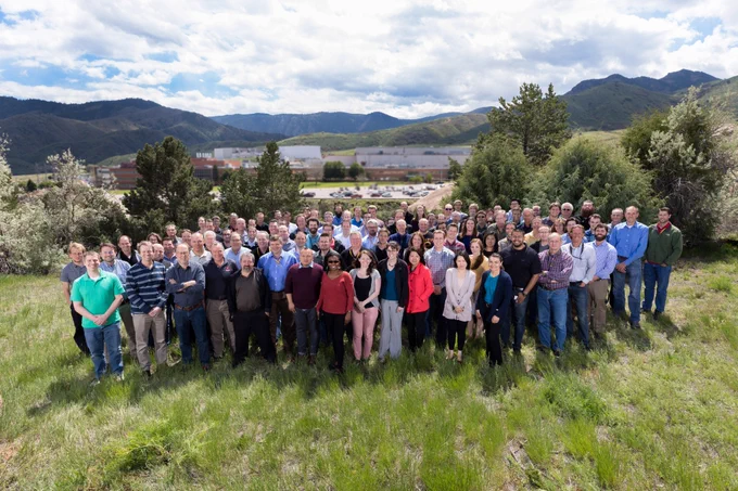 Posed group photo taken at Lockheed Martin shows around 100 people gathered in a grassy field, with buildings, trees, and mountains in the background.
