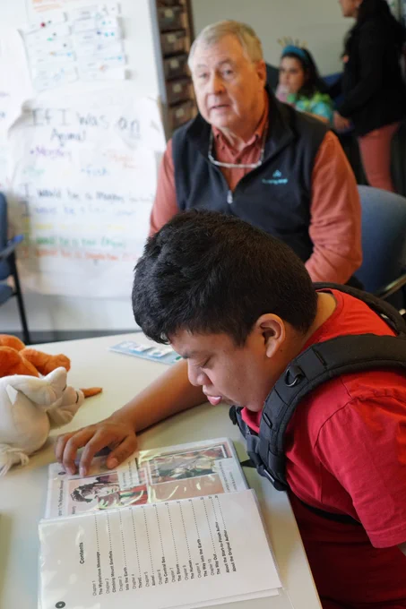A student in a red shirt looking at his Readtopia book with Don Johnston in the background.