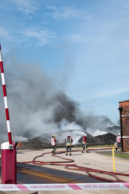 Three members of the Fire and Rescue Service using a hose to spray water on to large piles of burnt waste. There is a piece of tape in the foreground that says "FIRE DO NOT CROSS" and there is a small red brick building on the right.