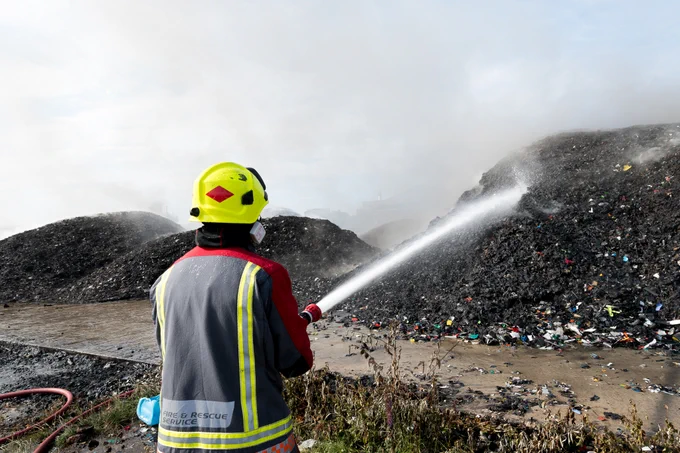 A member of the Fire and Rescue Service using a hose to spray water on to large piles of burnt waste.