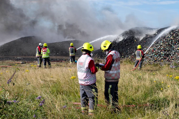 Three members of the Fire and Rescue Service using a house to spray water on to large piles of burnt waste. Two members of the Fire Service stand in the foreground talking and looking at a notepad.