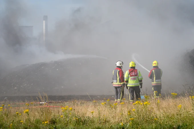 Three members of the Fire and Rescue Service using a hose to spray water on to large piles of burnt waste.