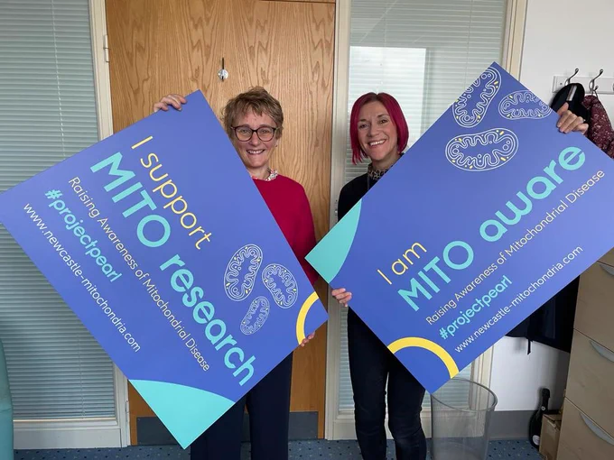 Amanda Temby and Lyndsey Butterworth from the Wellcome Centre for Mitochondrial Research holding the placards that they will take to the parliamentary event taking place to raise awareness of mitochondrial disease. The placards have a blue background with yellow and green writing that says "I support MITO research" and "I am MITO AWARE" followed by "Raising Awareness of Mitochondrial Disease #project pearl"