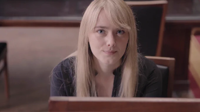 A young blonde woman sitting in a library looks straight at the camera with a gentle expression