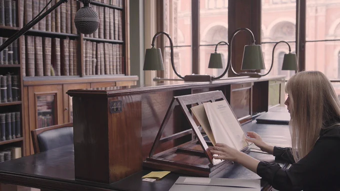 A reader with long blonde hair reads a magazine propped up on a lectern in a wood-panelled library overlooking a garden
