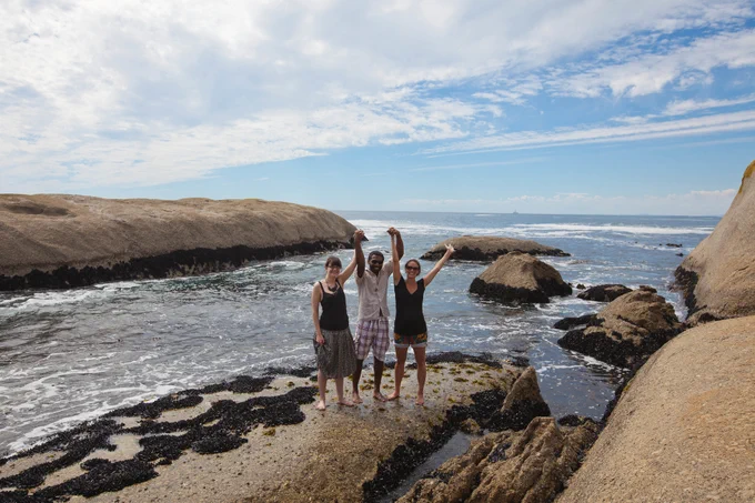Ariel, Kevin, and Caroline on a rock in the ocean holding hands in the air.