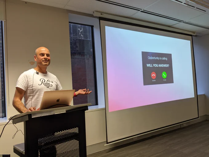 Tarun in front of his laptop, gesturing to his slideshow projected on the wall behind him. The slide reads, opportunity is calling. Will you answer? with decline or answer button below.  