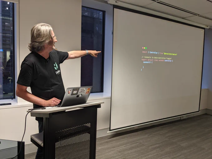 Lee in front of his laptop, pointing to his slideshow being projected on the wall behind him. The slide shows some code: import { controller } from "@hotwire/stimulus" 
// connects to data-controller="toggle"
export default class extends controller {
connect() {
   }
}