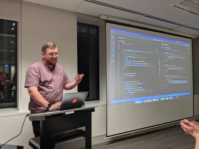 Michael in front of his laptop, smiling and gesturing toward the audience, with the projection of his screen on the wall behind him. The screen shows his solution to the protein translation Exercism challenge, though the code isn't readable as it's blurry. 