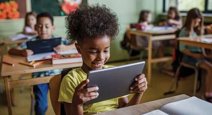 a classroom of young students with a close up on one student who is smiling while looking at a tablet