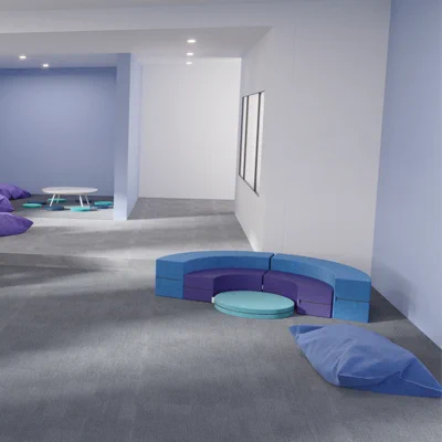 Purple foam-filled large cushions can be seen in the background of a corridor showing Beparta furniture. They are near a low white, writable table. There are light and dark teal low seat pads around the low table. In the foreground is amphitheatre-style foam seating in darker blue, purple and lighter blue. There are 3 tiers and each tier can be separated into 2 arcs. The lowest tier is a large round mat. Another large blue cushion is close to the viewer.