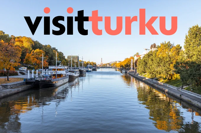 Joki kuvattuna suoraan edestä päin. Joen laitaan on ankkuroituna isoja aluksia ja joen rannalla lehtipuita syksyn väreissä. Taivas on vaaleansininen ja taivaan päällä on teksti "visitturku".