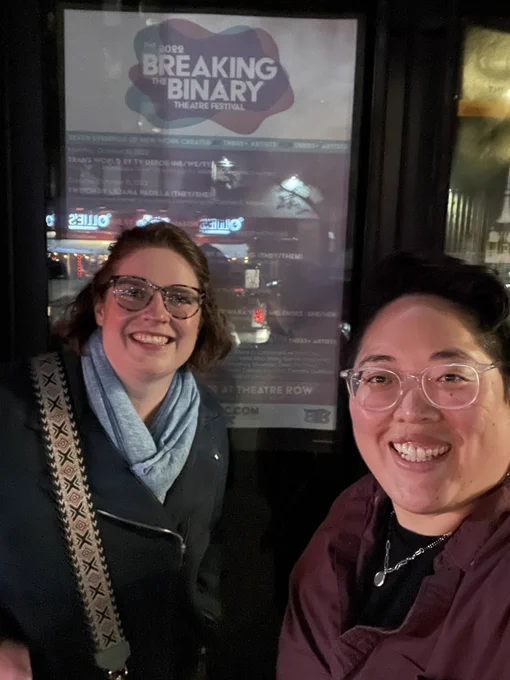 Photo of Meghan and Harrow smiling outside Theatre Row in front of the Breaking the Binary Theatre Festival poster. Meghan with shoulder-length curly brown hair wears tortoiseshell glasses, a light blue scarf, a blue jacket, and a bag with a black and brown patterned strap. Harrow, a non-binary Korean person with short black hair wears clear glasses, a maroon button-down, black t-shirt, and a silver necklace with a circle pendant.