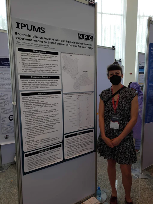 Picture of Maya standing next to the poster, also smiling while wearing a mask. 