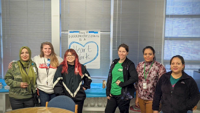 6 amazing Para's standing in a row Infront of a small poster that says "Being a Paraprofessional is a Heart Work"