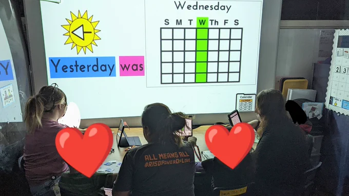 My para's, Ms. V, Ms. Ashlee and Ms. Mirna, running calendar. They are sitting at the table with their back to the camera with students sitting in between them. The board says "Yesterday was Wednesday"