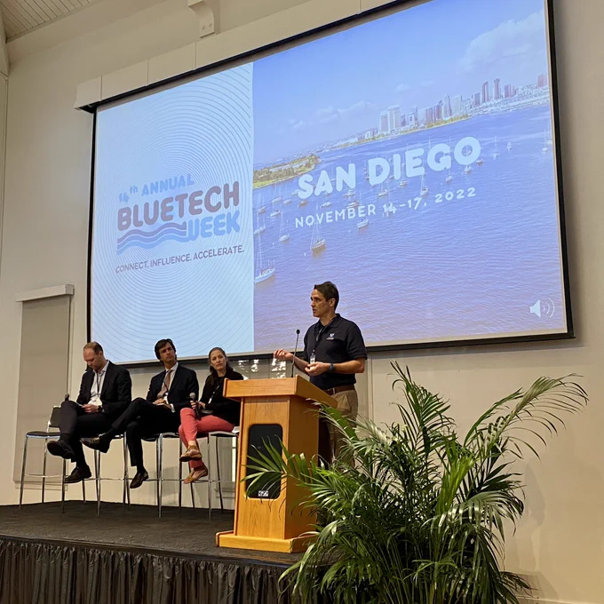 Stage with four people and projected background with text ‘San Diego BlueTech Week’
