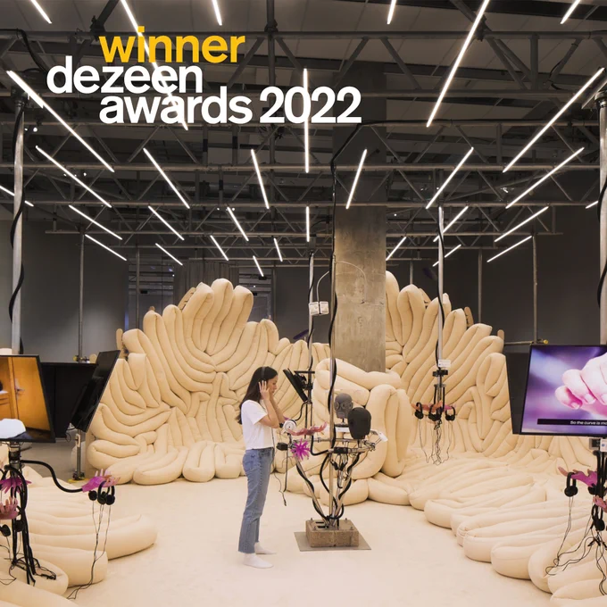 a woman stands in a room surrounded by beige sausage-like structures and screens, the text on top reads winner dezeen awards 2022