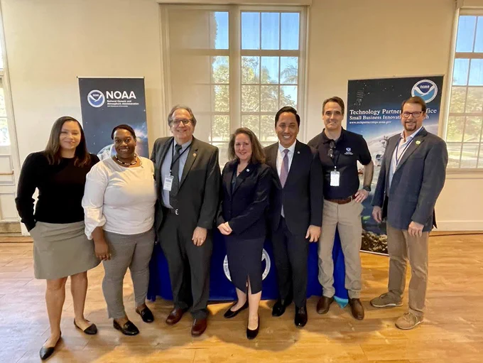 Seven people in business attire posing for photo in front of sign with the NOAA logo