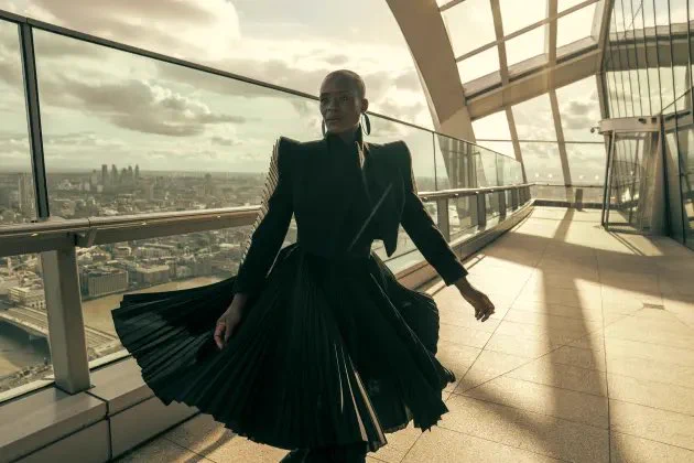 A Black woman with a shaved head is in mid turn. The long pleated skirt of her stylish black dress with accented shoulder flares around her. She is wearing large earrings.