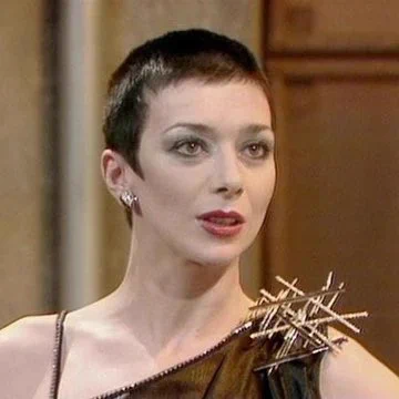 A white woman with very short hair wears a stylish off-the-shoulder black dress with a large angular brooch in the other shoulder. She is wearing earrings and impeccable makeup
