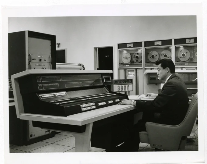 A white table console containing the black computer console, the Central Processing Unit of the mainframe is in front and off to the right in the background are banks of tape reading machines. The computer room has white tile and likely a raised floor for the wiring.